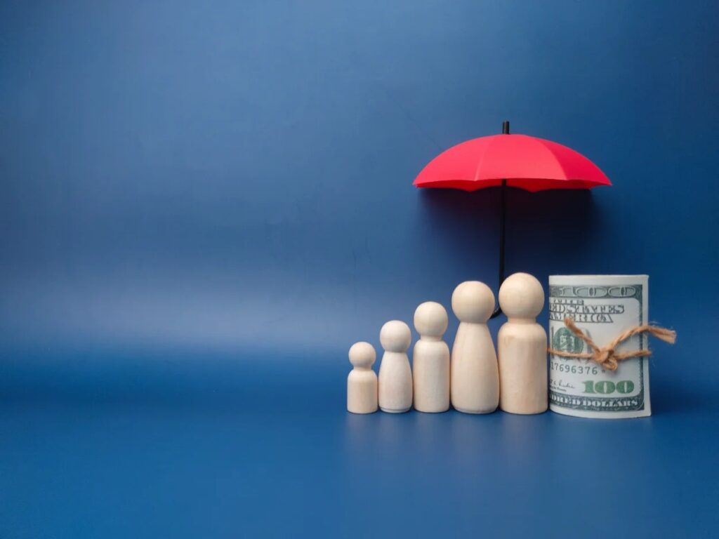 blue background with 5 wooden figures lined up from shortest to tallest with an umbrella overhead and a one-hundred dollar bill directly to the right